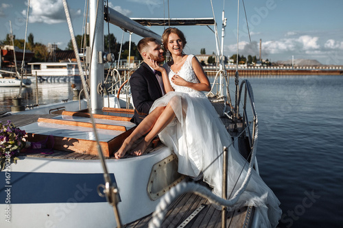 Wedding couple is hugging on a yacht. Beauty bride with groom. Beautiful model girl in white dress. Man in suit. Female and male portrait. Woman with lace veil. Cute lady and guy outdoors © svitlychnaja