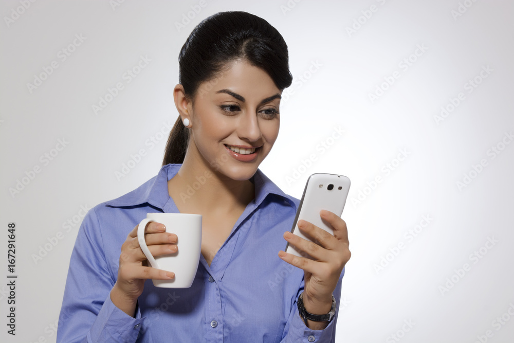 Businesswoman with coffee mug reading text message on smart phone against gray background Businesswoman with coffee mug reading text message on smart phone against gray background