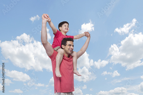 Happy father carrying son on shoulders against cloudy sky