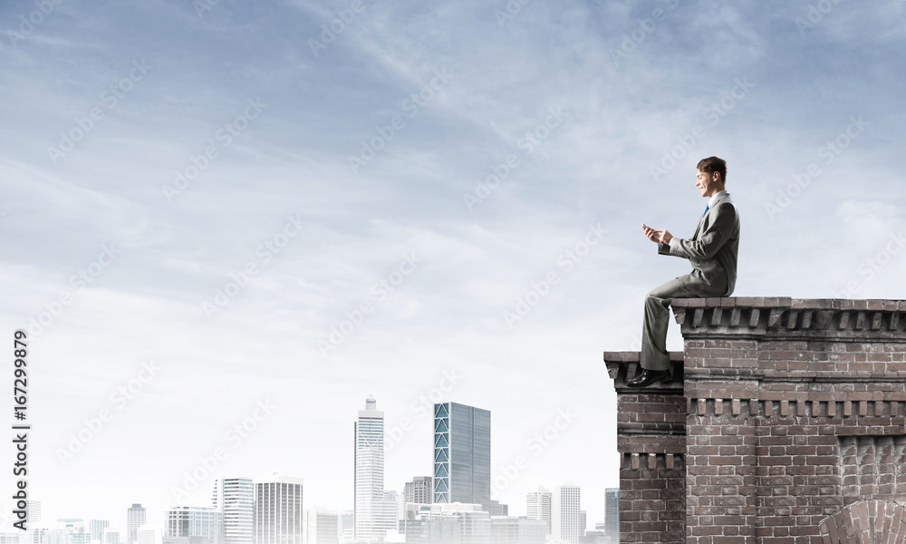 Man on roof edge send message with smartphone and cityscape at background Man on roof edge send message with smartphone and cityscape at background