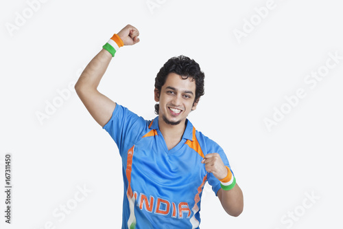 Portrait of young man in Indian cricket team jersey cheering up with clenched fists over white background 