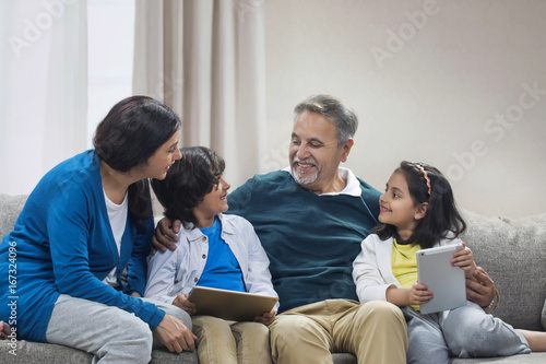 Happy grandparents and grandchildren using digital tablet sitting on sofa
