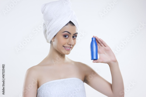 Portrait of confident woman wrapped in towel holding hair oil bottle against gray background