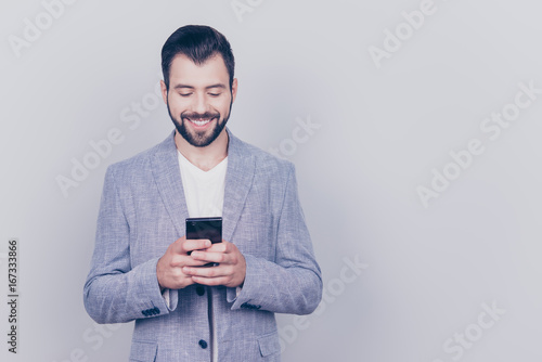 Young cheerful brunet bearded guy lawyer is standing on the pure light background and smiling, reading good news on internet at his mobile phone, wearing suit