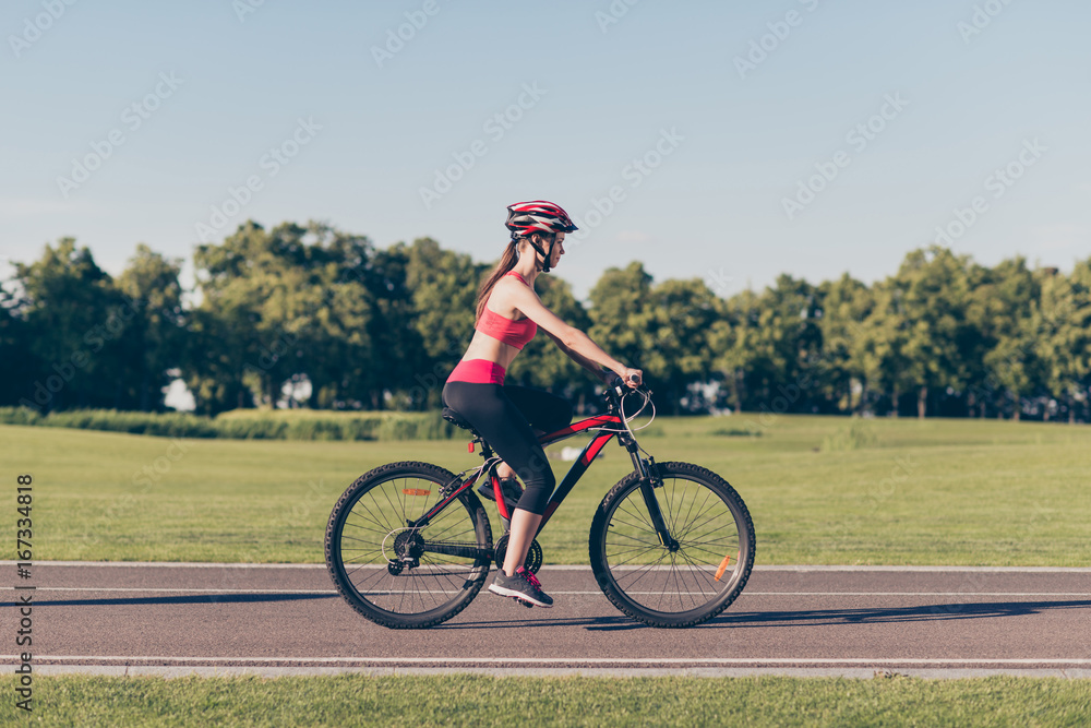Cute slim sport girl is riding modern bike outdoors in nice spring park, wearing helmet and trendy sport outfit, sneakers