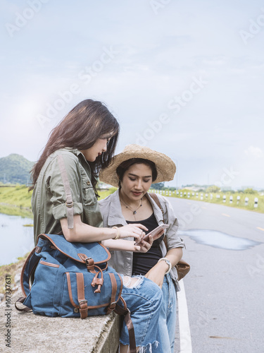 Two asian female traveller using smart phone for searching about location, young woman tourist sitting beside highway during planning about their trip. © Ketsada