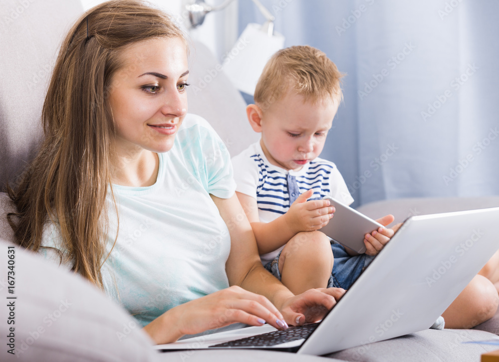 Smiling mom is productively working behind laptop while kid playing on tablet