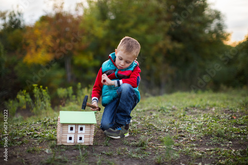 A small boy in an autumn forest collects a wooden house designer, holding a hammer in his hand. © @VMStock