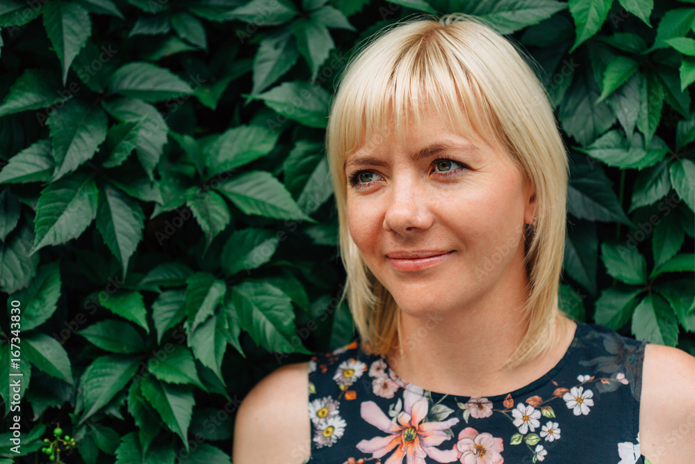 Cute young blonde stylish woman in pretty blouse in flowers over green grape leaves background. Beautiful smiling girl