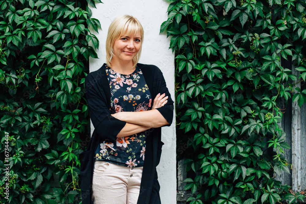 Cute young blonde beautiful stylish woman in pretty blouse in flowers and black cardigan over green grape leaves background.