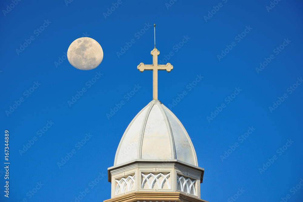 The white church roof has a cross, with the moon and the sky as the background.