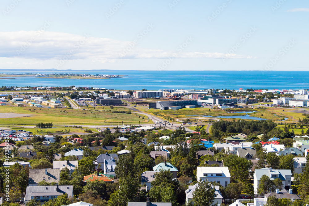 Aerial view on capital of Iceland - city of Reykjavik - ocean bay, port, green meadows, roadways, vehicles, streets, houses, traffic, roofing - on sunny day.