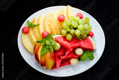 classic summer Italian food. Beautifully decorated fruit platter with watermelon, melon, apricot and grapes on a white plate, dark background, top view