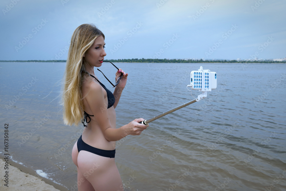 Sexy young blonde wearing sunglasses doing selfie on the beach. Happy cute  girl with a attractive ass. Caucasian model take skin care therapy. Concept  of selfie-stick. Cool color Stock Photo | Adobe