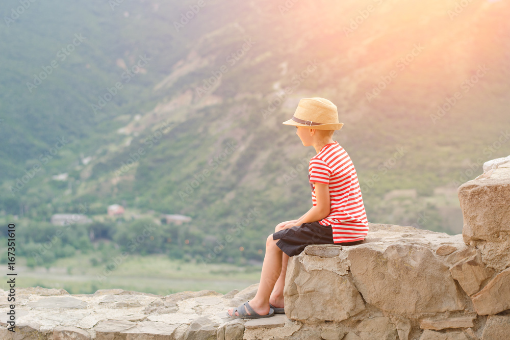 Boy in a hat sitting on stones on a background of green mountains