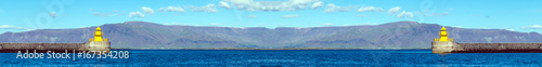 Yellow lighthouse in ocean bay against blue sky and mountains on horizon. Copy space. Leaderboard. Wide panoramic image.