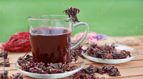 Roselle juice in cup and dry roselle on wooden plank against green grass background.