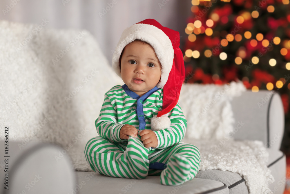 Cute little baby in Santa hat sitting on sofa in decorated for Christmas room Cute little baby in Santa hat sitting on sofa in decorated for Christmas room