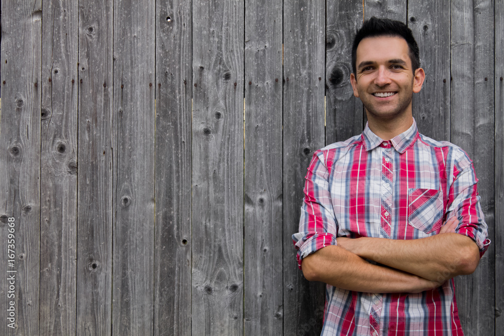 Obraz premium Portrait of handsome young man in shirt keeping arms crossed and smiling while standing against wooden background.