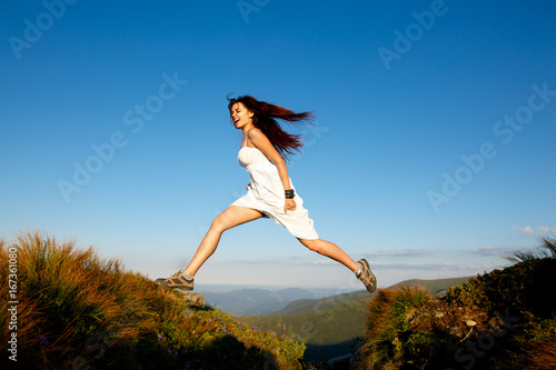 Young woman in white dress running in the mountains. Woman jumping on mountain peak rock. Beautiful girl looking happy and smiling