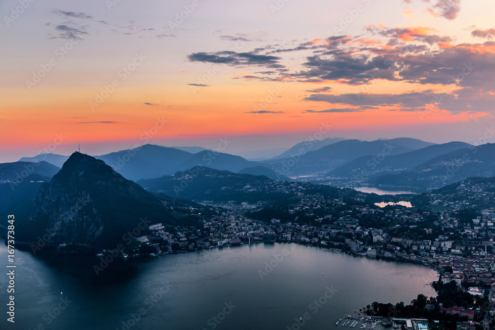 Aerial view of the lake Lugano surrounded by mountains and evening city Lugano on during dramatic sunset, Switzerland, Alps. Travel Aerial view of the lake Lugano surrounded by mountains and evening city Lugano on during dramatic sunset, Switzerland, Alps. Travel