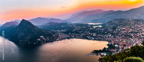 Aerial view of the lake Lugano surrounded by mountains and evening city Lugano on during dramatic sunset, Switzerland, Alps. Travel photo