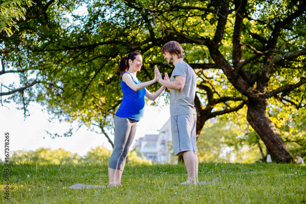 Fototapeta premium Pregnant couple workout outdoor - restraining palm to palm while standing on yoga mat in park. Parenthood, sport and healthy life concept.