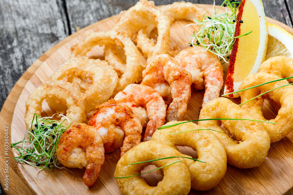 Seafood platter with deep fried squid rings, shrimp and onion rings decorated with lemon on cutting board on wooden background. Mediterranean appetizers. Top view Seafood platter with deep fried squid rings, shrimp and onion rings decorated with lemon on cutting board on wooden background. Mediterranean appetizers. Top view