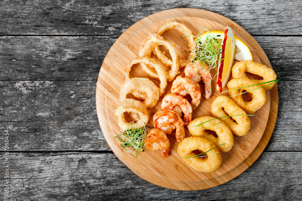 Seafood platter with deep fried squid rings, shrimp and onion rings decorated with lemon on cutting board on wooden background. Mediterranean appetizers. Top view Seafood platter with deep fried squid rings, shrimp and onion rings decorated with lemon on cutting board on wooden background. Mediterranean appetizers. Top view