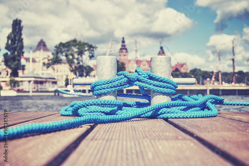 Color toned picture of a cleat with blue rope on a wooden pier, Szczecin waterfront in distance, Poland.