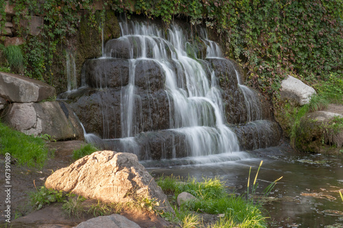 Small waterfall in Alexandria park  Belaya Tserkov  Ukraine. Shot on a sunny hot summer day. Golden setting sun light on the rock near the river with the waterfall in the background