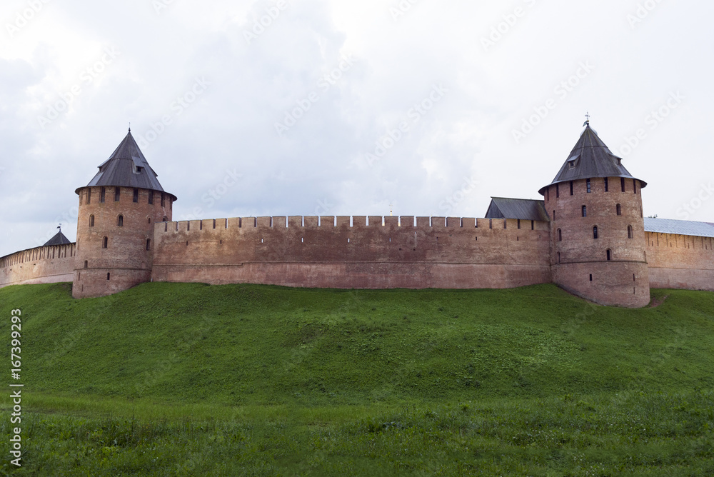 medieval fortification wall with towers of red brick, the Novgorod Kremlin, the green grass, rain clouds