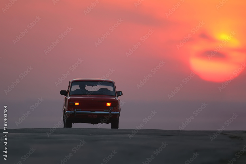 Old red car driving on the road towards sunrise