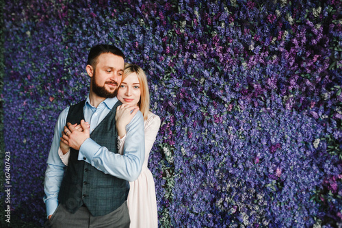 Portrait of a loving young woman embracing man from behind. A man in a waistcoat and shirt and a woman hugging from behind in a pink dress holding hands against bright flowers. photo