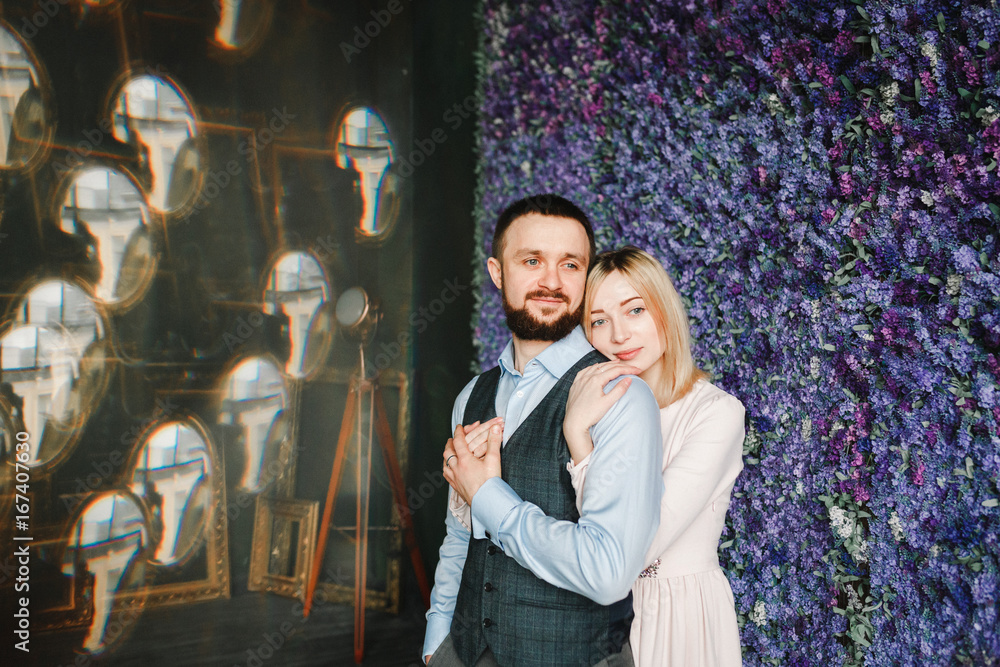 Portrait of a loving young woman embracing man from behind. A man in a waistcoat and shirt and a woman hugging from behind in a pink dress holding hands against bright flowers. Portrait of a loving young woman embracing man from behind. A man in a waistcoat and shirt and a woman hugging from behind in a pink dress holding hands against bright flowers.