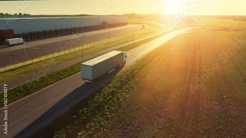 Aerial Follow Shot of White Semi Truck with Cargo Trailer Attached Moving Through Industrial Warehouse, Rural Area. Sun Shines and the Sky Are Blue. Shot on RED EPIC-W 8K Helium Cinema Camera.