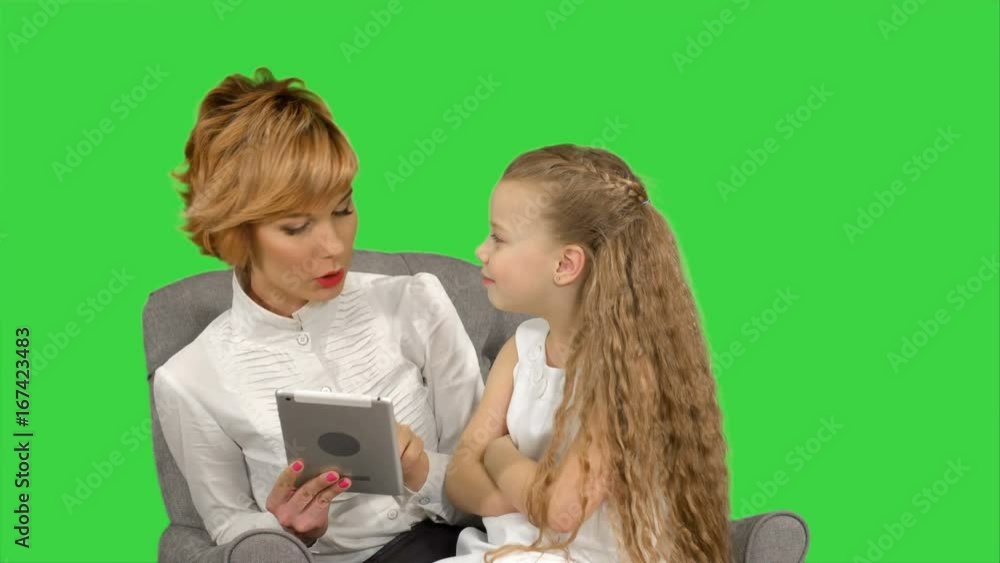 Young mother and her daughter sit together and look at the tablet on a Green Screen, Chroma Key Young mother and her daughter sit together and look at the tablet on a Green Screen, Chroma Key
