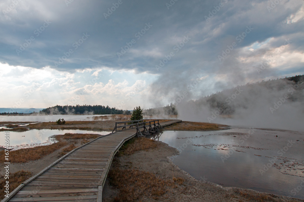 Elevated wooden boardwalk under heavy rain  clouds going past Black Warrior Springs and Tangled Creek and overlooking Hot Lake in the Lower Geyser Basin in Yellowstone National Park in Wyoming USA
