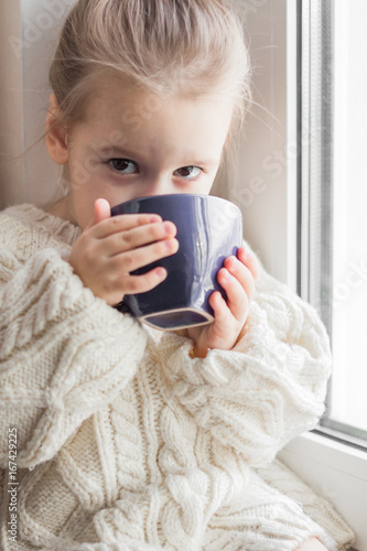 A little girl in the white knitted sweater sits on a window and drinks from a cup