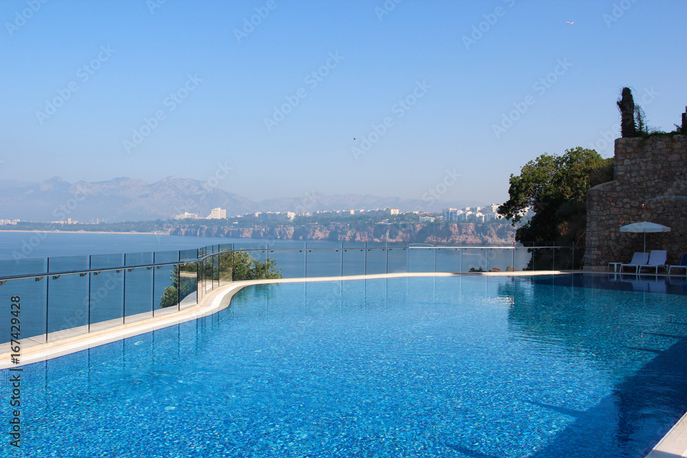 A view of the pool and the sea with turquoise clear water with a mountain range on the horizon and the line of the Mediterranean coast with the resort town in distance. 
