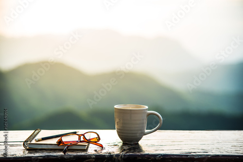  A coffee cup, eyeglasses and a note book on wooden table with sunrise and mountain  bokeh background. A start of new day with hot beverage. Business concept.