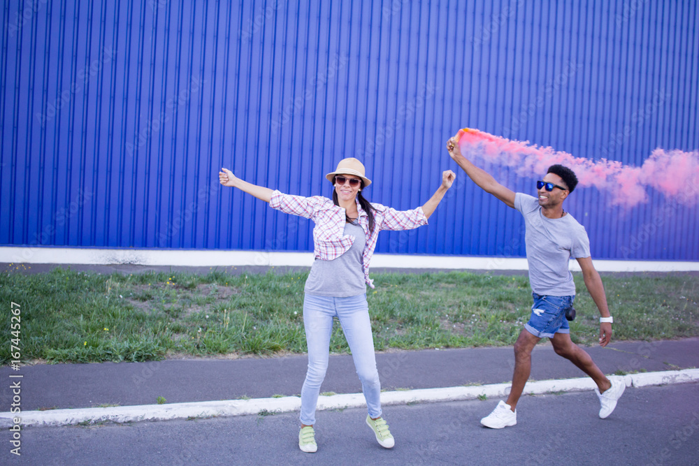 Young couple riding on skateboards with color smoke bomb, boy and girl in casual clothes having good time, pink and blue colors  