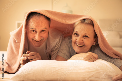 Senior couple lie together under the covers. Older couple lie on bed and looking at camera.