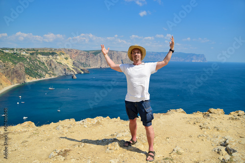 A man stands on top of the mountain against the sea and blue sky. Spread his arms to the side, looking at the camera and smiling. Crimea, Russia. Horizontal orientation © alexbard