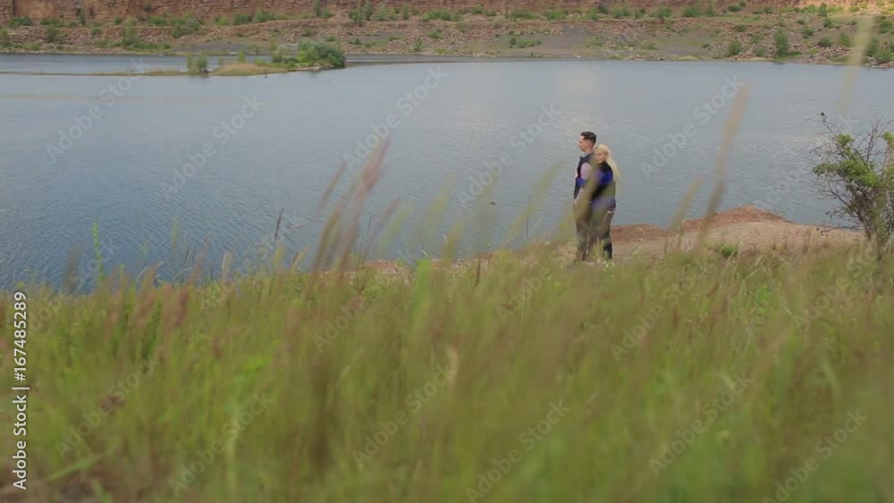 Girl hugging her husband from behind standing at a beautiful lake. Wide shot. Copy space. In the foreground of swaying grass. Girl hugging her husband from behind standing at a beautiful lake. Wide shot. Copy space. In the foreground of swaying grass.