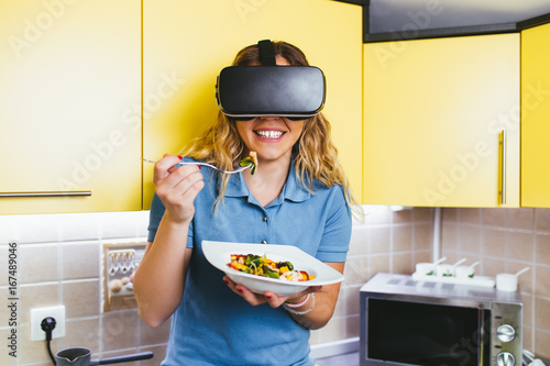 Beautiful attractive young woman eating healthy vegetable meal with VR glasses on her. Healthy lifestyle concept. 