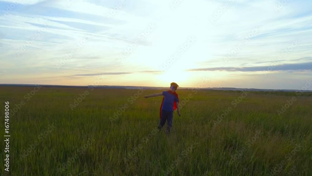 A boy in a superman costume runs across the green field at sunset A boy in a superman costume runs across the green field at sunset
