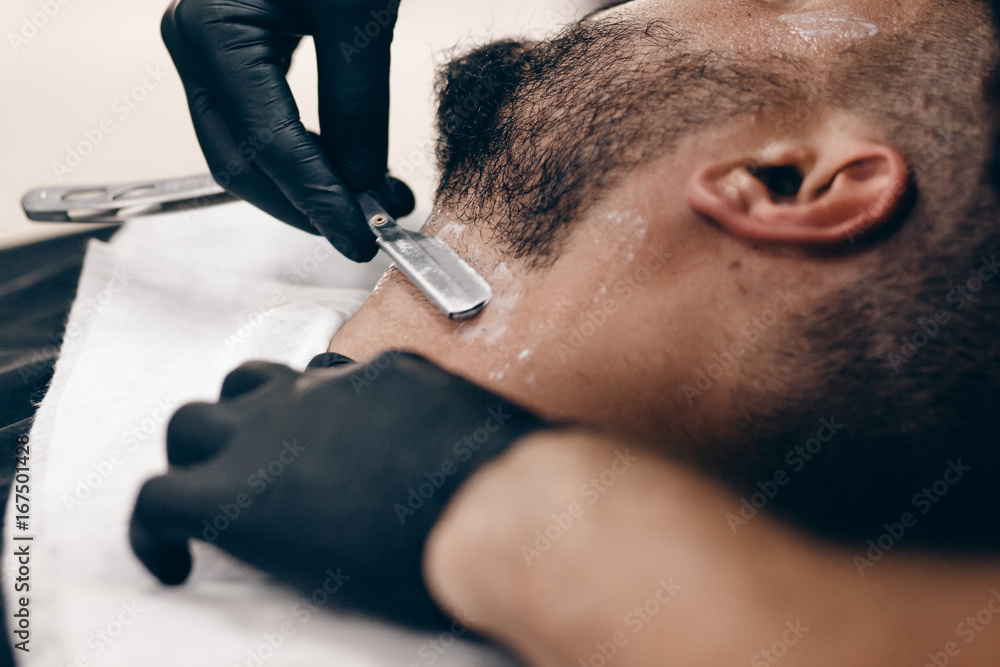 A man with a beard a client in a barbershop hairdresser on a shaving with a dangerous razor. A man with a beard a client in a barbershop hairdresser on a shaving with a dangerous razor.