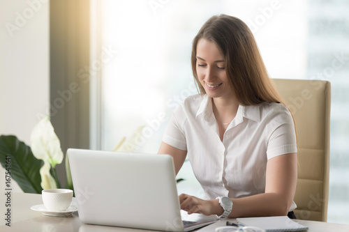Smiling businesswoman using laptop computer in office, happy employee communicating online at workplace, secretary typing email, chatting via internet, planning daily work with easy business software