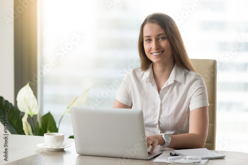 Attractive businesswoman working on laptop and smiling for camera in workplace, successful confident executive manager sitting at office desk, young e-business owner posing at work, headshot portrait